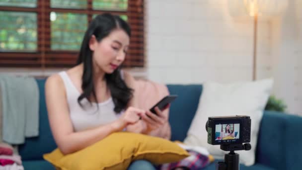 アジアのファッションインフルエンサーデザイナーの女性がカメラストリーミングを使用してライブで服ビジネスを販売し モバイル電話チャットを使用してオンラインで電子商取引放送自宅のリビングルームでソファの上で顧客に答える — ストック動画