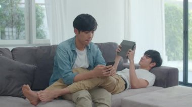 Modern evde cep telefonu ve tablet kullanan genç eşcinsel lgbtq çifti. Asyalı aşık erkek mutlu, rahatla, gül ve eğlenceli teknoloji internette oyun oynarken oturma odası konseptinde kanepede uzan..