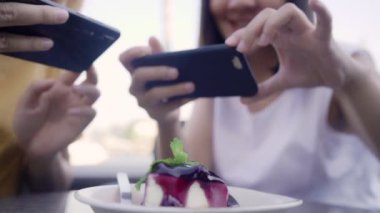 Neşeli genç Asyalı arkadaş kahve dükkanında fotoğraf yemek ve kek yerken telefon kullanıyor. Lokantada ya da kafede birlikte iki neşeli ve çekici Asyalı bayan. Tatil aktivitesi, ya da modern yaşam tarzı konsepti.
