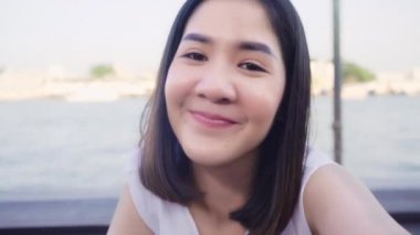 Blogger Asyalı arkadaş canlısı kadın etkileyicisi kafede kameraya bakıyor. Genç bayan, mutlu bir şekilde rahatlayın. Şehirdeki nehrin yakınındaki sosyal medya tatil seyahatinde video kaydını kullanarak rahatlayın..