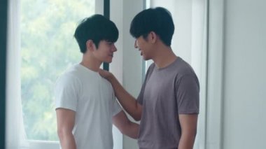 Asyalı eşcinsel çift evlerinin penceresinin önünde durup sarılıyorlar. Genç Asyalı Lgbtq + erkekler mutlu öpüşürler birlikte dinlenirler modern evdeki oturma odasında romantik zaman geçirirler..
