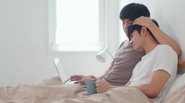 Asyalı eşcinsel çift bilgisayar kullanıyor ve modern evde kahve içiyor. Genç Asyalı aşık erkek mutlu. Uyandıktan sonra birlikte dinlenin. Sabahları yatak odasında uzanmış film seyredin.