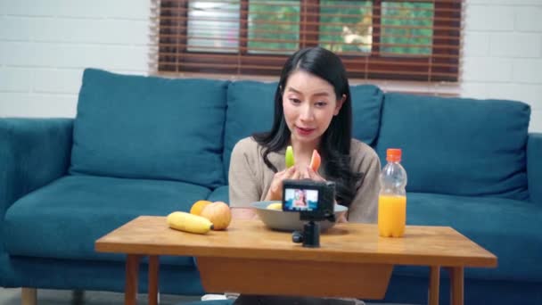 アジアの影響力のある女性は オンライン放送のために自宅のリビングルームでソファの上に果物の健康的な食べ物を食べることの利点についてのビデオをストリーミングや記録するカメラを使用して — ストック動画