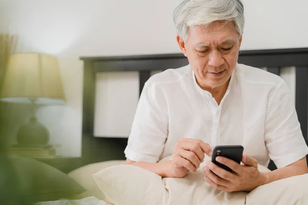Asiatiska äldre män som använder mobiltelefon hemma. Asiatiska Senior kinesiska manliga sökning information om hur man god hälsa på internet när du ligger på sängen i sovrummet hemma på morgonen konceptet. — Stockfoto