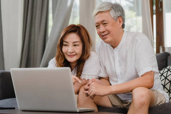 아시아 선배 부부 가 집에서 노트북을 사용하고 있습니다. 아시아 출신의 중국인 조부모들은 가정에서 소파에 누워 소셜 미디어를 확인하기 위해 인터넷 서핑을 한다.. — 스톡 사진