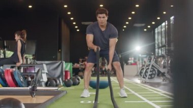 Genç atlet Asyalı adam, spor salonundaki halatlarla mücadele ederek yağ yakma egzersizinin bir parçası olarak ipleri dalgalarla hareket ettiriyor. Sporcu eğlencesi, sağlıklı yaşam tarzı konsepti..