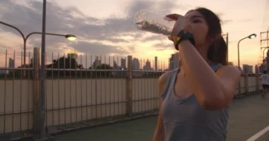 Güzel genç Asyalı atlet bayan şehir ortamında koştuktan sonra yorgun hissettiği için içme suyu egzersizi yapıyor. Japon genç kız sabah erken saatlerde yürüyüş köprüsünde spor giysileri giyerek spor yapıyor..