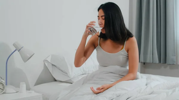 Malata malata femmina indiana soffre di insonnia. Asiatica giovane donna che prende antidolorifico medicina per alleviare il mal di testa e bere bicchiere d'acqua seduto sul letto nella sua camera da letto a casa al mattino. — Foto Stock