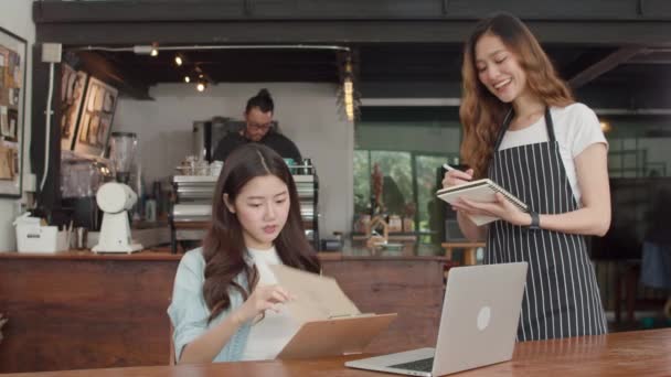 都会のカフェで若い客の女の子から注文メニューを受け取るノート付きの陽気なアジアの女性のウエイトレス コーヒーショップでラップトップで働く若いアジア人のフリーランス女性 所有者小規模事業コンセプト — ストック動画