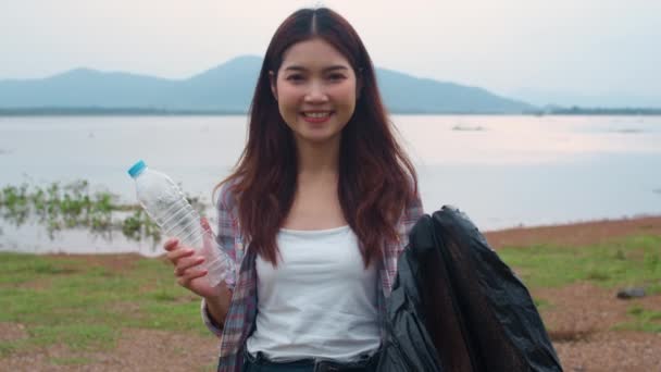 年轻的亚洲女性志愿者的肖像有助于保持大自然的清洁 他们在海滩上拿着塑料瓶废物和黑色垃圾袋 环境保护及污染问题的概念 — 图库视频影像