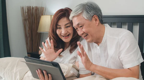 Asiatisches Seniorenpaar mit Tablet zu Hause. Asiatische ältere chinesische Großeltern, Videotelefonie im Gespräch mit Enkelkindern der Familie, während sie morgens im Schlafzimmer zu Hause auf dem Bett liegen. — Stockfoto