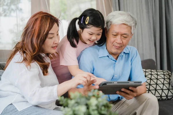 Asian grandparents and granddaughter using tablet at home. Senior Chinese, grandpa and grandma happy spend family time relax with young girl checking social media, lying on sofa in living room concept — Stock Photo, Image