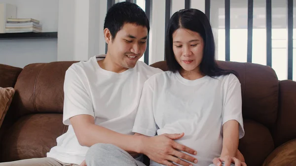 Joven asiática Embarazada pareja hombre tocar su esposa vientre hablando con su hijo. Mamá y papá se sienten felices sonriendo tranquilos mientras cuidan al bebé, el embarazo acostado en el sofá en la sala de estar en el concepto de casa. —  Fotos de Stock