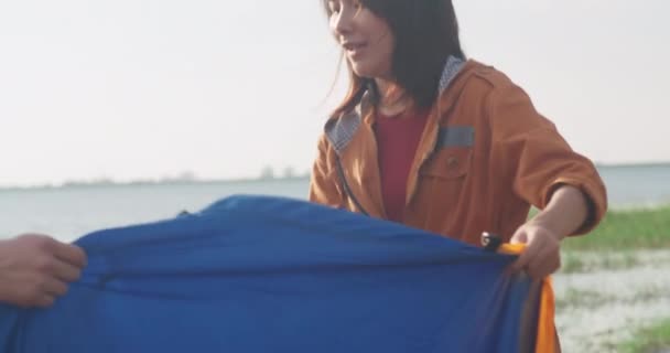 若いアジアのキャンパーカップルは 海辺の近くにテントキャンプ用具を屋外に設置しました 男性と女性の旅行はキャンプ場で夏の日に楽しんでいます 野外活動 冒険旅行 または休暇 — ストック動画