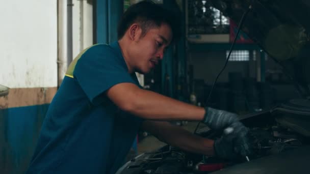 Profesionální automechanik šroubuje detaily automobilového motoru na zdvižený automobil na opravárenské čerpací stanici. Dovedný Asiat v uniformě opravuje auto v mechanické garáži v noci. 4k zpomalený záběr.