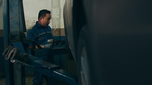 修理サービスステーションで解除された自動車のタイヤを変更する専門的な車の整備士 夜間の作業場でのホイールアライメント作業 均一な固定車の熟練したアジア人の男 4Kスローモーションショット — ストック動画