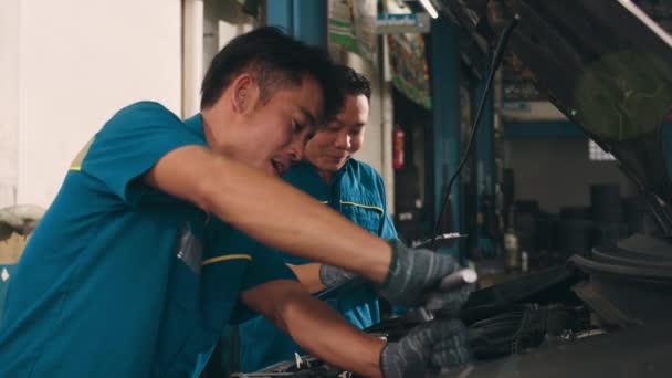 修理サービスステーションで解除された車のエンジンの2つのプロの車の整備士のネジ詳細 ガレージの夜に均一な固定車の熟練したアジア人の男 車のサービスの概念 スローモーション — ストック動画