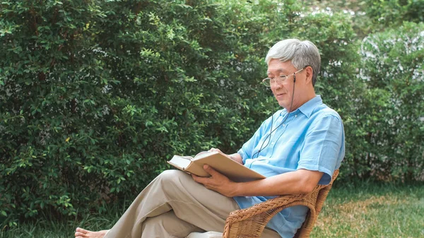 Aasialaiset vanhemmat miehet rentoutuvat kotona. Aasian vanhempi kiinalainen mies nauttia lepoajasta kulumista lasit lukea kirjoja, kun makaa puutarhassa kotona aamulla käsite. — kuvapankkivalokuva