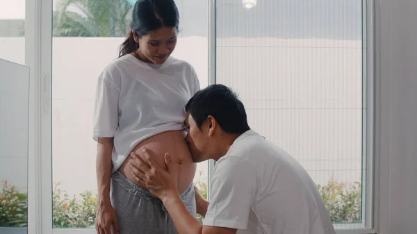 Młody azjatycki mężczyzna w ciąży całujący żonę w brzuch rozmawiający ze swoim dzieckiem. Mama i tata czują się szczęśliwi uśmiechając się spokojnie, dbając o dziecko, ciąża w pobliżu okna w salonie koncepcja domu. — Zdjęcie stockowe