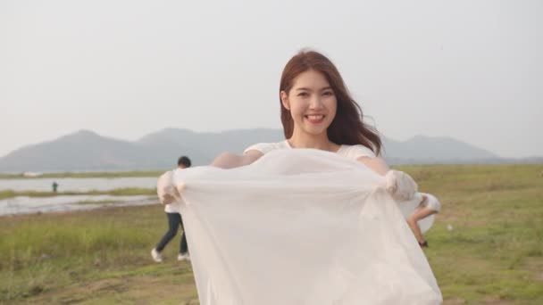 年轻的亚洲女性志愿者的肖像有助于保持自然的清洁 看着镜头 带着白色的垃圾袋在海滩上微笑 环境保护及污染问题的概念 — 图库视频影像