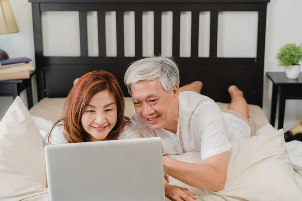 Asiática pareja de ancianos utilizando el ordenador portátil en casa. Asiático Senior abuelos chinos, marido y esposa feliz después de despertar, viendo la película acostado en la cama en el dormitorio en casa en el concepto de la mañana. — Foto de Stock