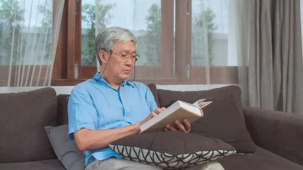 Aasialaiset vanhemmat miehet rentoutuvat kotona. Aasian Senior kiinalainen mies nauttia lepoajasta lukea kirjaa, kun makaa sohvalla olohuoneessa kotona käsite . — kuvapankkivalokuva