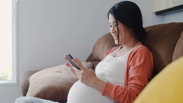 Młoda kobieta w ciąży azjatyckiej za pomocą telefonu komórkowego wyszukiwania ciąży informacji. Mama uczucie szczęśliwy uśmiech pozytywny i spokojny podczas dbać o swoje dziecko leżące na kanapie w salonie koncepcja domu. — Zdjęcie stockowe