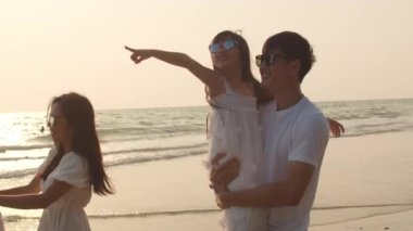 Asyalı genç mutlu aile akşamları sahilde tatilin tadını çıkarıyor. Baba, anne ve çocuk gün batımında deniz kenarında yürürken rahatlar. Hayat tarzı seyahat tatil yaz tatili konsepti. 4k ağır çekim.