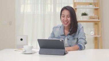 Asya 'da serbest çalışan kadınlar günlük kıyafetlerini tablet kullanarak giyerler iş yerindeki müşterilerle evlerinde video konferans yaparlar. Mutlu Asyalı genç kız, rahat ol masada otur ve internette işini yap..