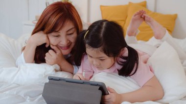Tablet kullanan Asyalı büyükanne torununa masal okuyor. Yaşlı Çinli, büyükanne mutlu. Yatmadan önce genç bir kızla rahatla. Yatak odasında, gece konseptinde..