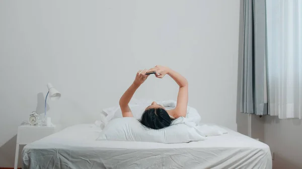 Mujer asiática joven usando smartphone comprobando las redes sociales sintiéndose feliz sonriendo mientras está acostada en la cama después de despertarse por la mañana, hermosa dama hispana atractiva sonriendo relajarse en el dormitorio en casa. —  Fotos de Stock