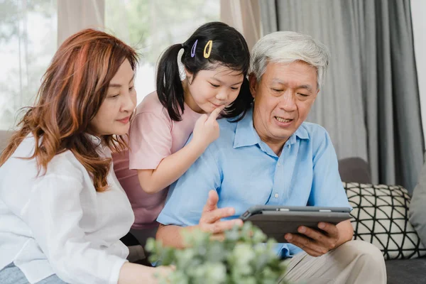 Asian grandparents and granddaughter using tablet at home. Senior Chinese, grandpa and grandma happy spend family time relax with young girl checking social media, lying on sofa in living room concept — Stock Photo, Image