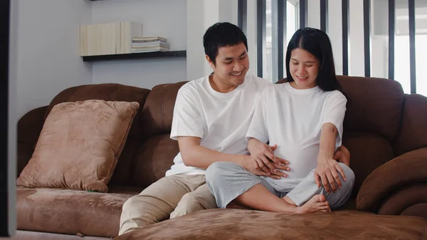 Unga asiatiska gravida par man röra hans fru mage talar med sitt barn. Mamma och pappa känner sig glada leende fridfull medan ta hand baby, graviditet ligger på soffan i vardagsrummet hemma koncept. — Stockfoto