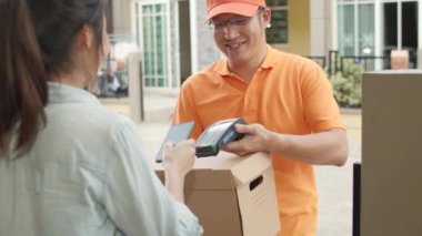 Genç posta kuryesi, paketleri müşteriye göndermek için elinde tutuyor, Asyalı kadınlar ise dışarıda paket teslim almak için cep telefonuna QR kodu ile imza atıyor ve ödeme yapıyor. 4k Ağır çekim.
