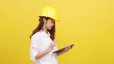 Genç Asyalı kadın mühendis pozitif ifadeli dijital tablet kullanıyor, günlük kıyafetler giyip sarı arka plandan kameraya bakıyor. Mutlu, mutlu, mutlu kadın başarıya sevinir..