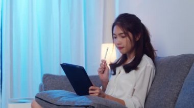 Tablet kullanan Asyalı iş kadını, gece evde oturma odasında çalışırken iş arkadaşlarıyla görüntülü görüşme planı hakkında konuşuyor. Kişisel izolasyon, sosyal uzaklık, koronavirüsü önlemek için karantina.