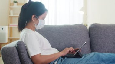 Asyalı serbest çalışan, koruyucu maske takan, evden oturma odasına tablet kullanan bir iş kadını. Sosyal uzaklık evde ve kendi kendine karantina süresi, koronavirüs konsepti....