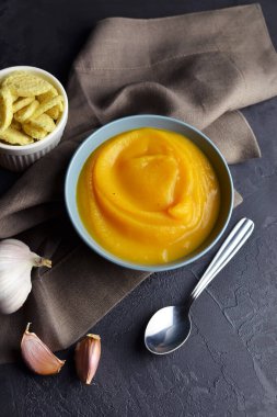 Kabak kremalı çorba, kızarmış ekmek ve seramik tabakta taze sarımsak. Siyah arka planda kaşık ve keten peçete.