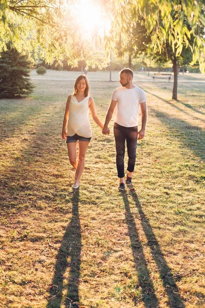 husband with pregnant wife holding hands and walking in park at sunny day