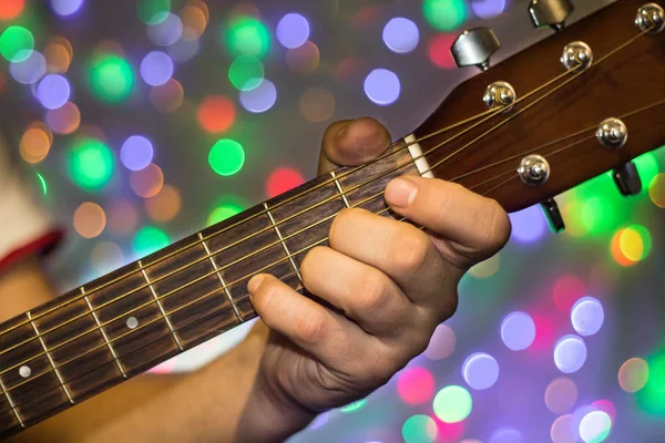 Homme jouant de la guitare acoustique. Doigts rapprochés sur le cou de la guitare contre les lumières bokeh floues de Noël sur fond — Photo