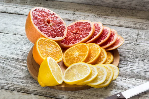 Limones frescos en rodajas, pomelos, naranjas en una tabla de cortar de madera con un cuchillo — Foto de Stock