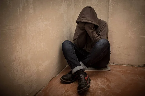 Foto van wanhopige jonge drugsverslaafde dragen kap en zitten alleen in hoek — Stockfoto