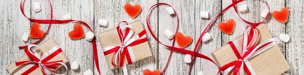 Banner of Valentines Day candy hearts marshmallows and box of gifts in craft paper over white wooden background.