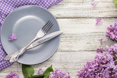 beyaz ahşap arka planda leylak çiçekleriyle süslenmiş çatal ve bıçakla döşenmiş bahar masası.