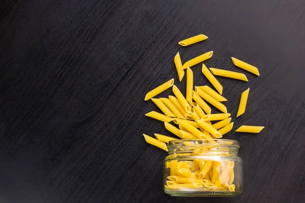 Glass jar filled with dry penne pasta and scattered around on black background — Stock Photo, Image