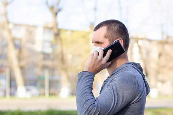 Caucasian man in medical mask on his face calling by phone. Man in protective mask is protected from virus in street outdoor.