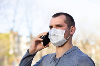 Yüzünde tıbbi maske olan beyaz bir adam telefonla arıyor. Koruyucu maskeli adam dışarıda virüsten korunuyor..