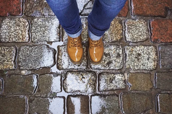 Look down at the feet in yellow shoes and blue jeans on the background of a wet old paving stone after the rain.