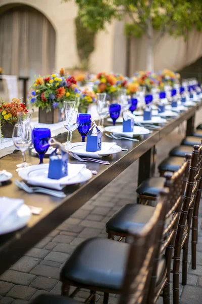 オレンジと黄色の花で飾られたパティオの結婚式のテーブルセット ミッションスタイルのパティオの庭で素朴なテーブルの上の青とクリスタル風のガラス — ストック写真