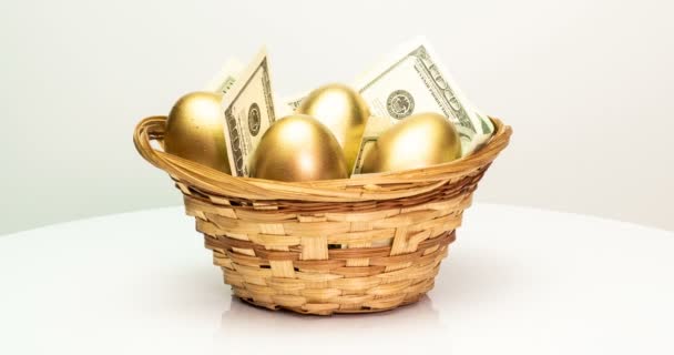 Золотые яйца и доллары в корзине на белом фоне. Золотые яйца крупным планом, съедобные, весенние, инвестиционные и пенсионные концепции. Вращение камеры — стоковое видео